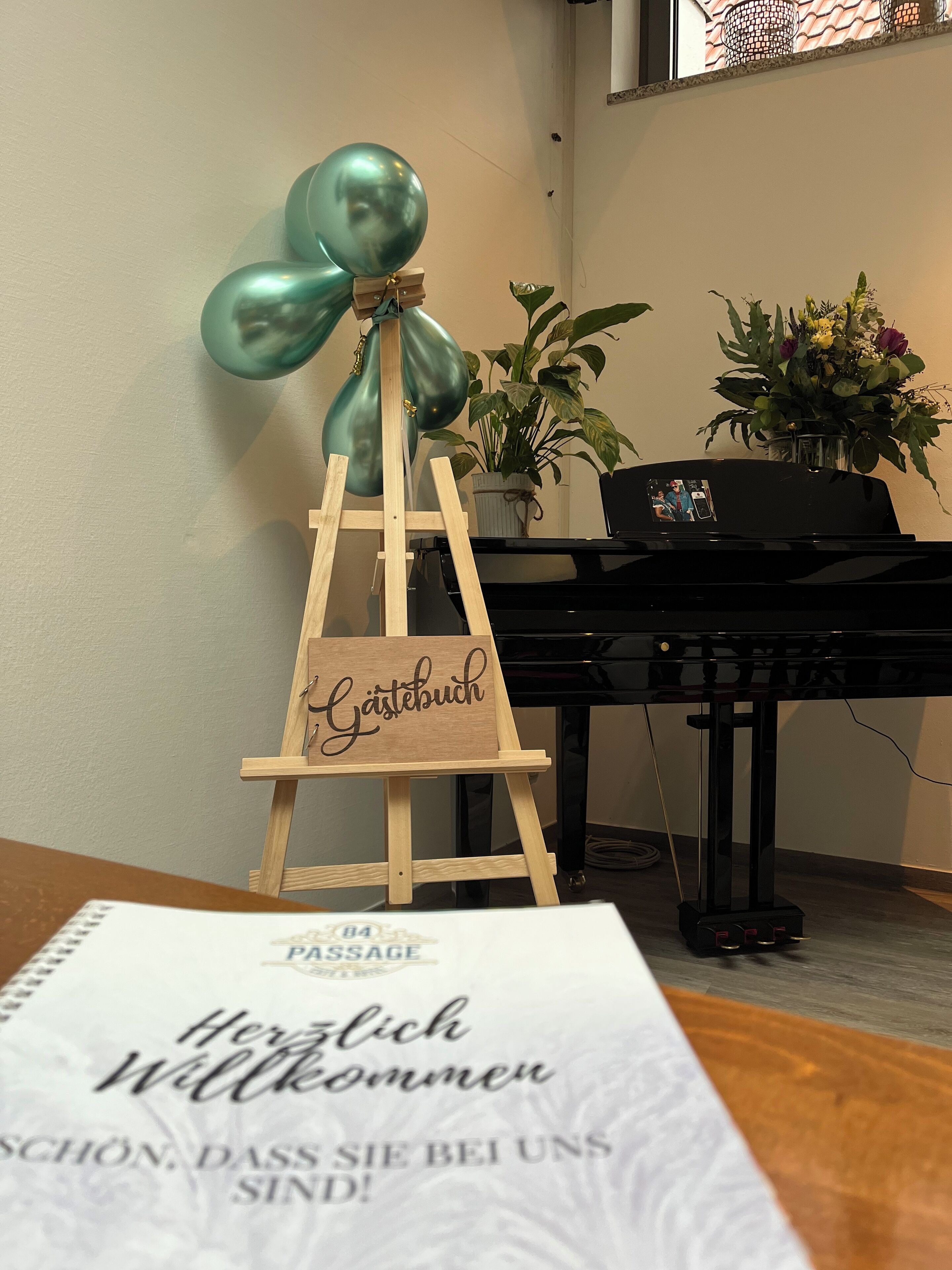 A wooden easel with a sign reading "Gästebuch" and a teal balloon tied to it. In the background, there is a piano and a potted plant with flowers.