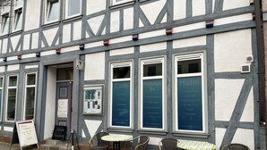 Um edifício tradicional de estrutura de madeira com uma placa de café, mesas ao ar livre com cadeiras de vime e uma pequena mesa.