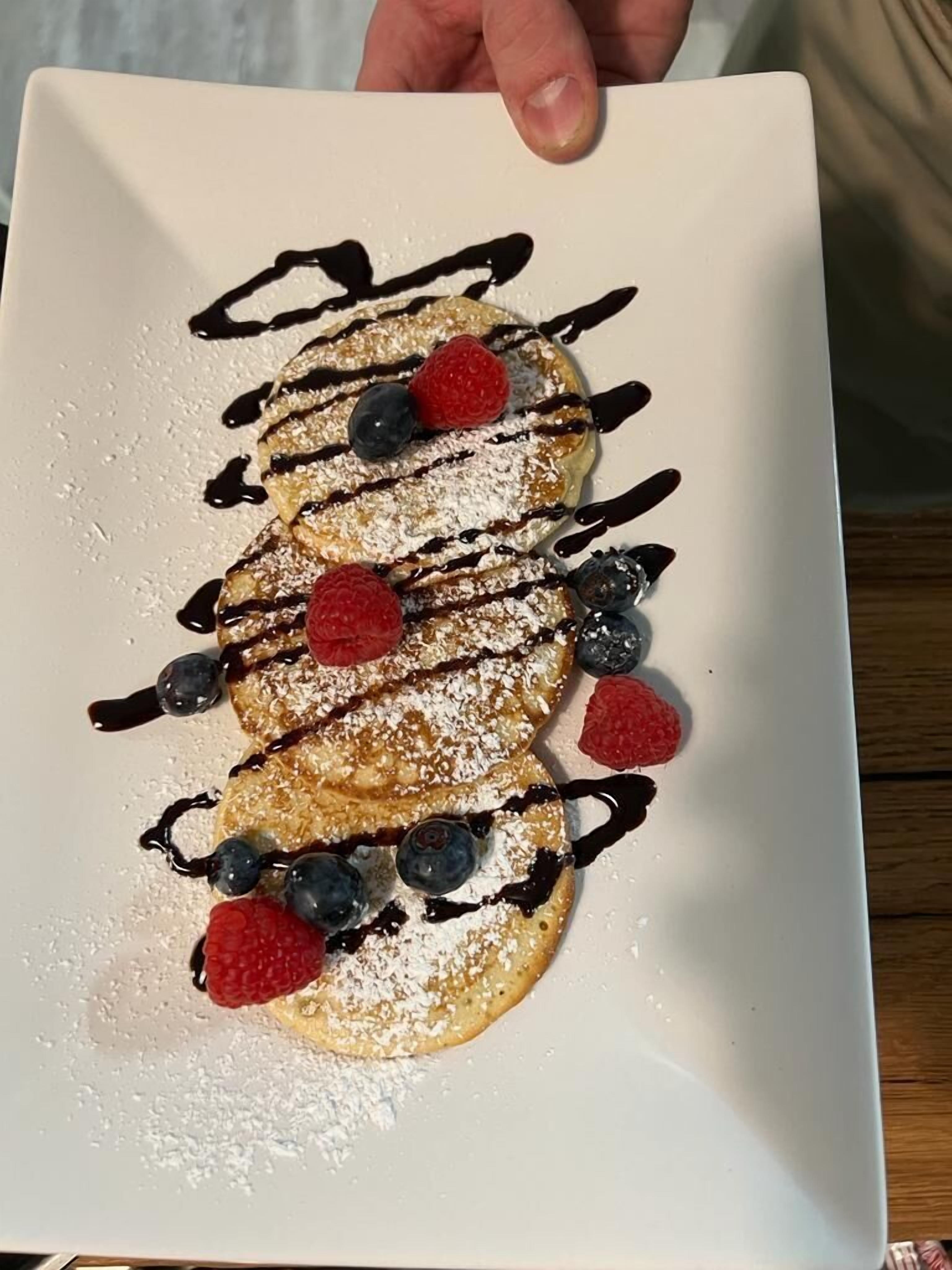 A square plate with a stack of pancakes topped with powdered sugar, blueberries, raspberries, and chocolate drizzle.