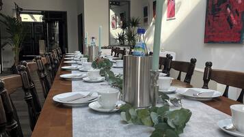 A long dining table set for a meal with white plates, cups, and cutlery, surrounded by chairs.