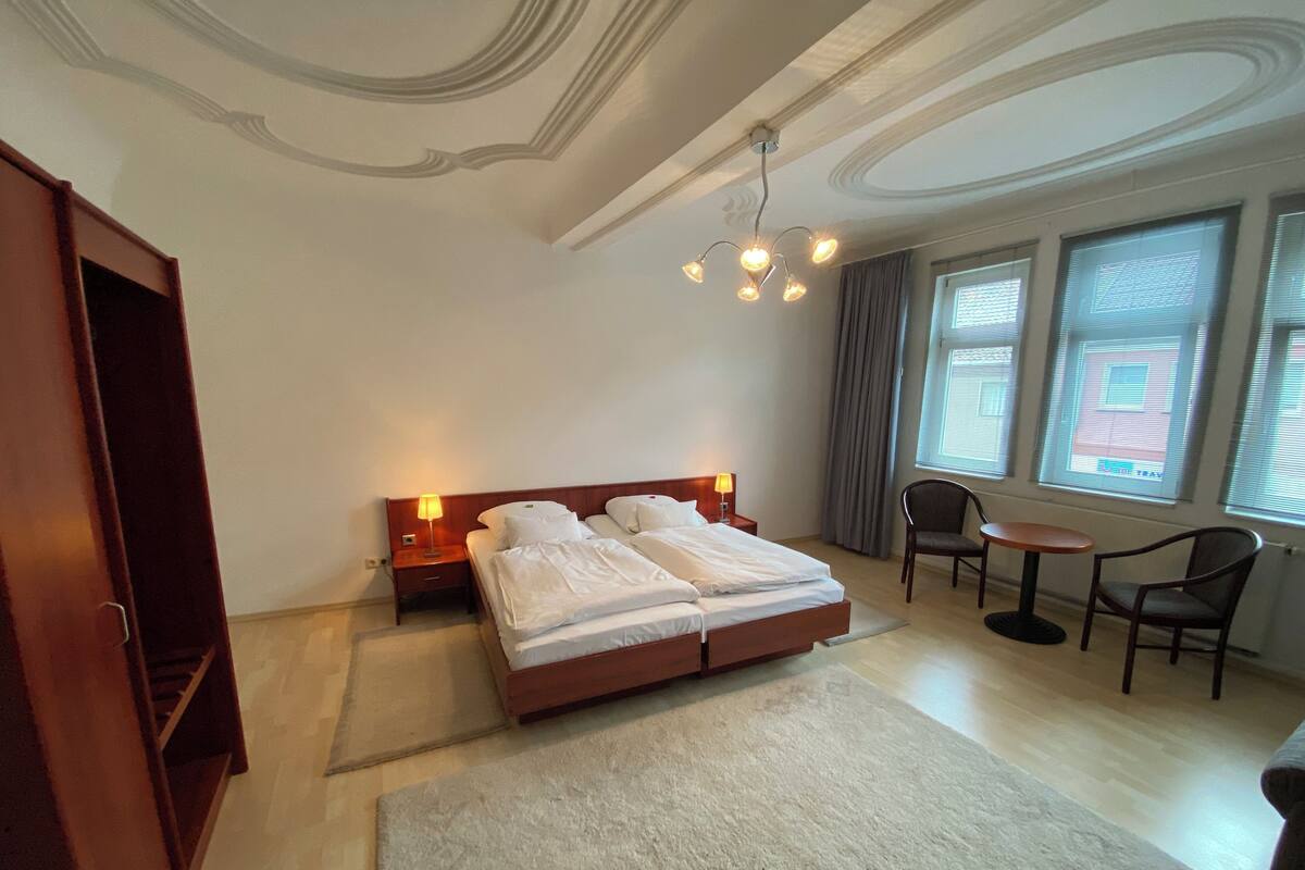 Ein Hotelzimmer mit einem Doppelbett, zwei Nachttischen mit Lampen, einem kleinen runden Tisch mit zwei Stühlen, einem großen Fenster mit Vorhängen und einem Holzschrank.