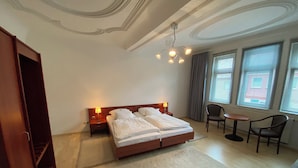Quarto de hotel com cama de casal, duas mesas de cabeceira com luminárias, uma mesa redonda pequena com duas cadeiras, uma janela grande com cortinas e um guarda-roupa de madeira.