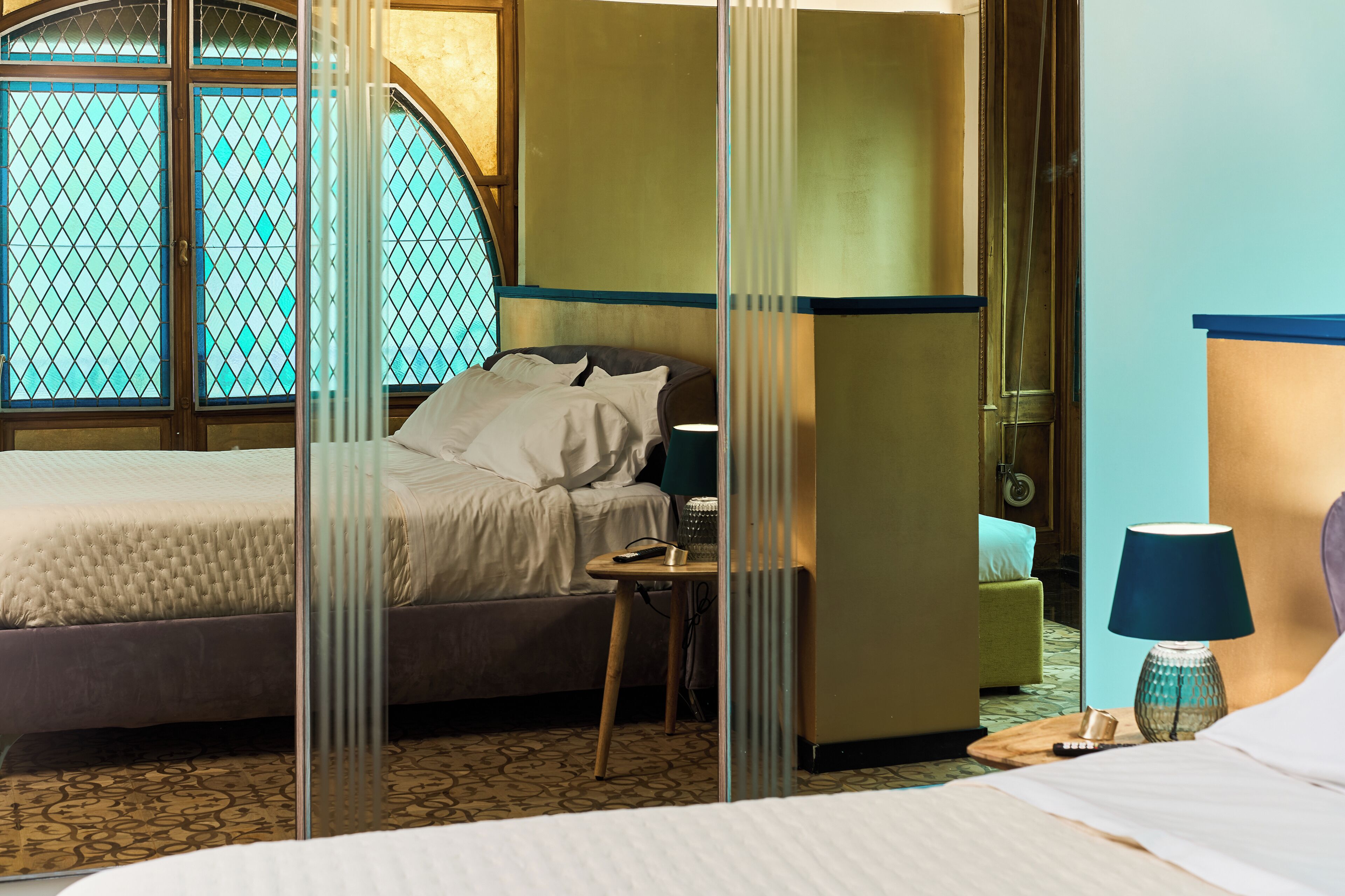 Ein Hotelzimmer mit Bett, Nachttisch, Lampe und Blick durch ein Fenster mit dekorativem Sichtschutz.