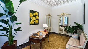 A room with a wooden table, a sofa, a large potted plant, a painting of yellow roses, and a cabinet with gold accents. - GLAM TIME (Genoa)