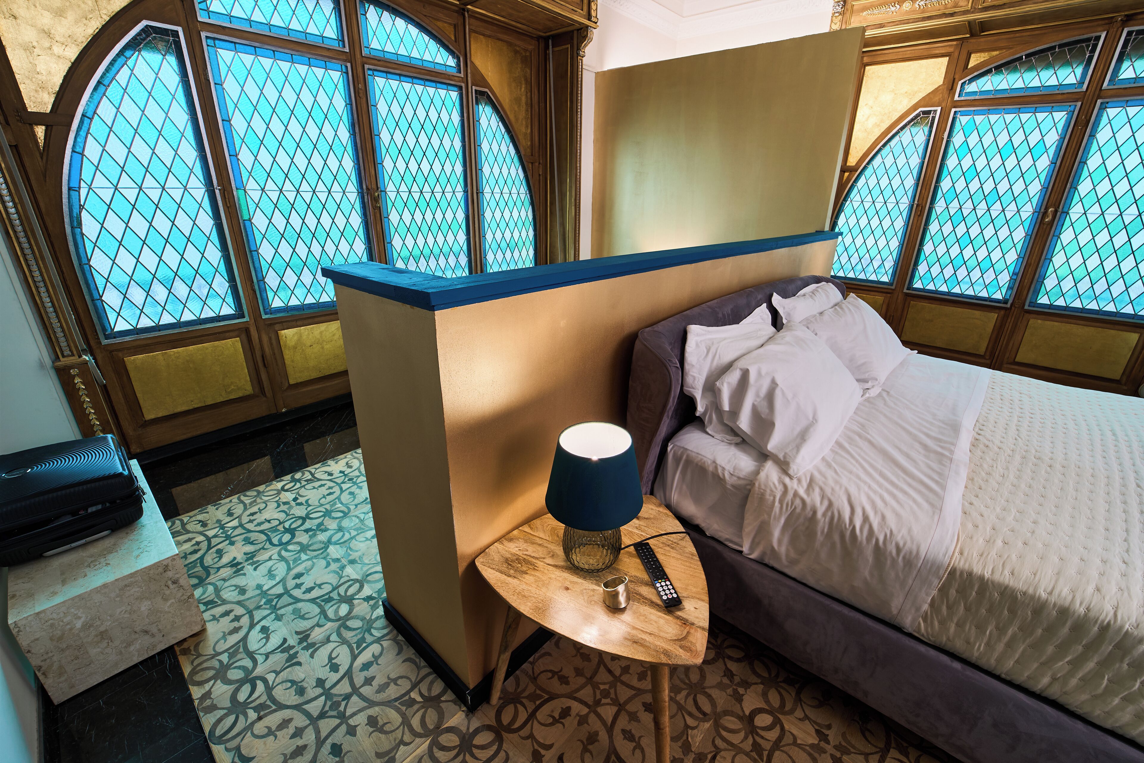 A room with a bed, a small table with a lamp, a remote control, and a suitcase.