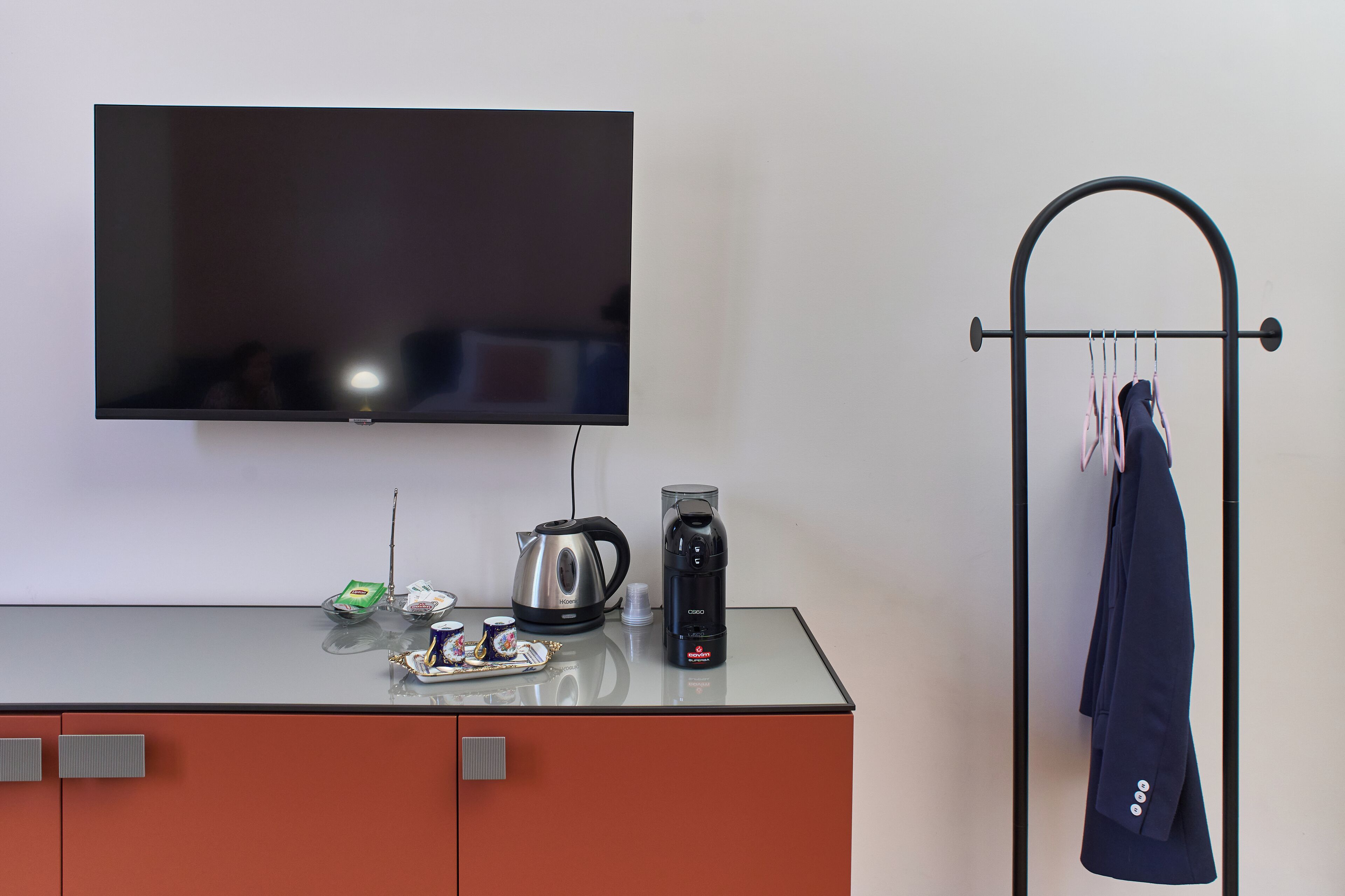 A modern room with a flat-screen TV mounted on the wall, a coffee maker, a kettle, and a coat rack with a coat hanging on it.