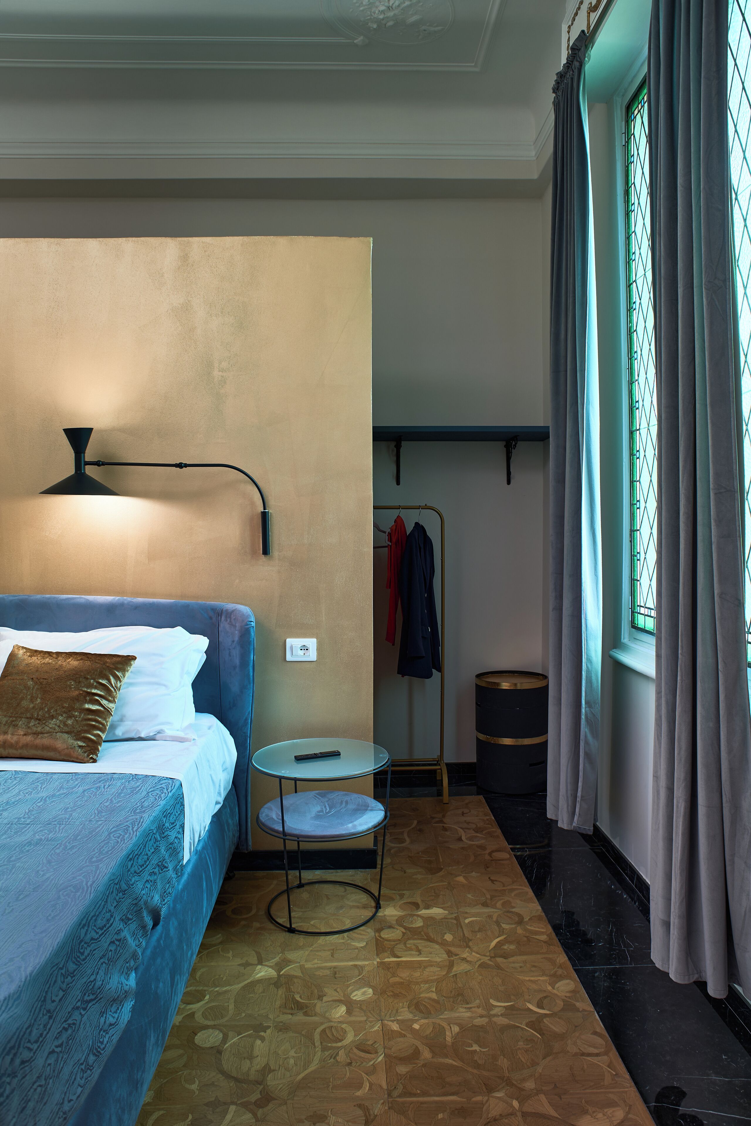 Quarto de hotel com cama, uma mesa redonda, um banquinho, um cabideiro e janela com cortinas.