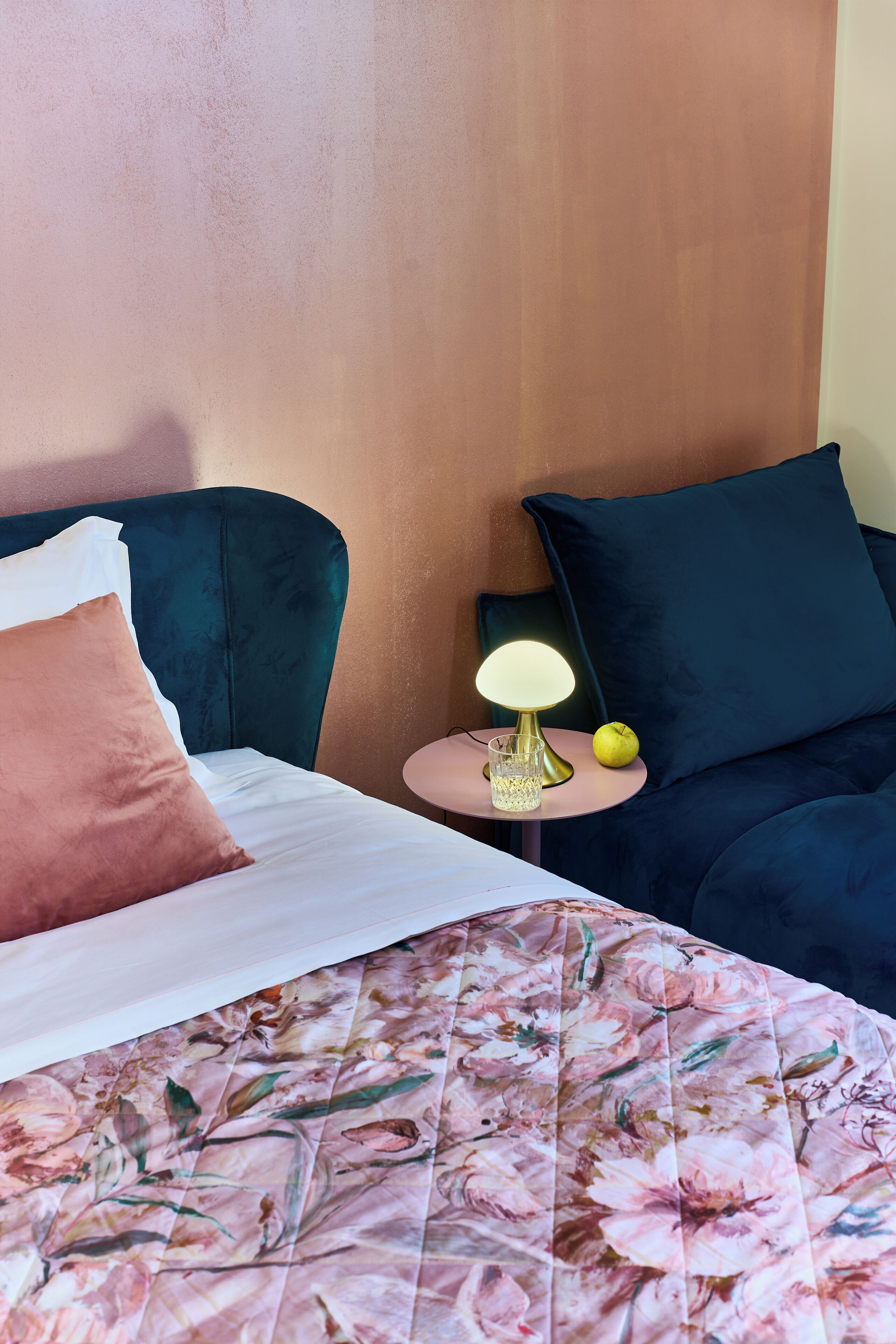 Ein ordentlich bezogenes Bett mit Blumenmuster, eine Nachttischlampe und ein kleiner Tisch mit Glas und Apfel.