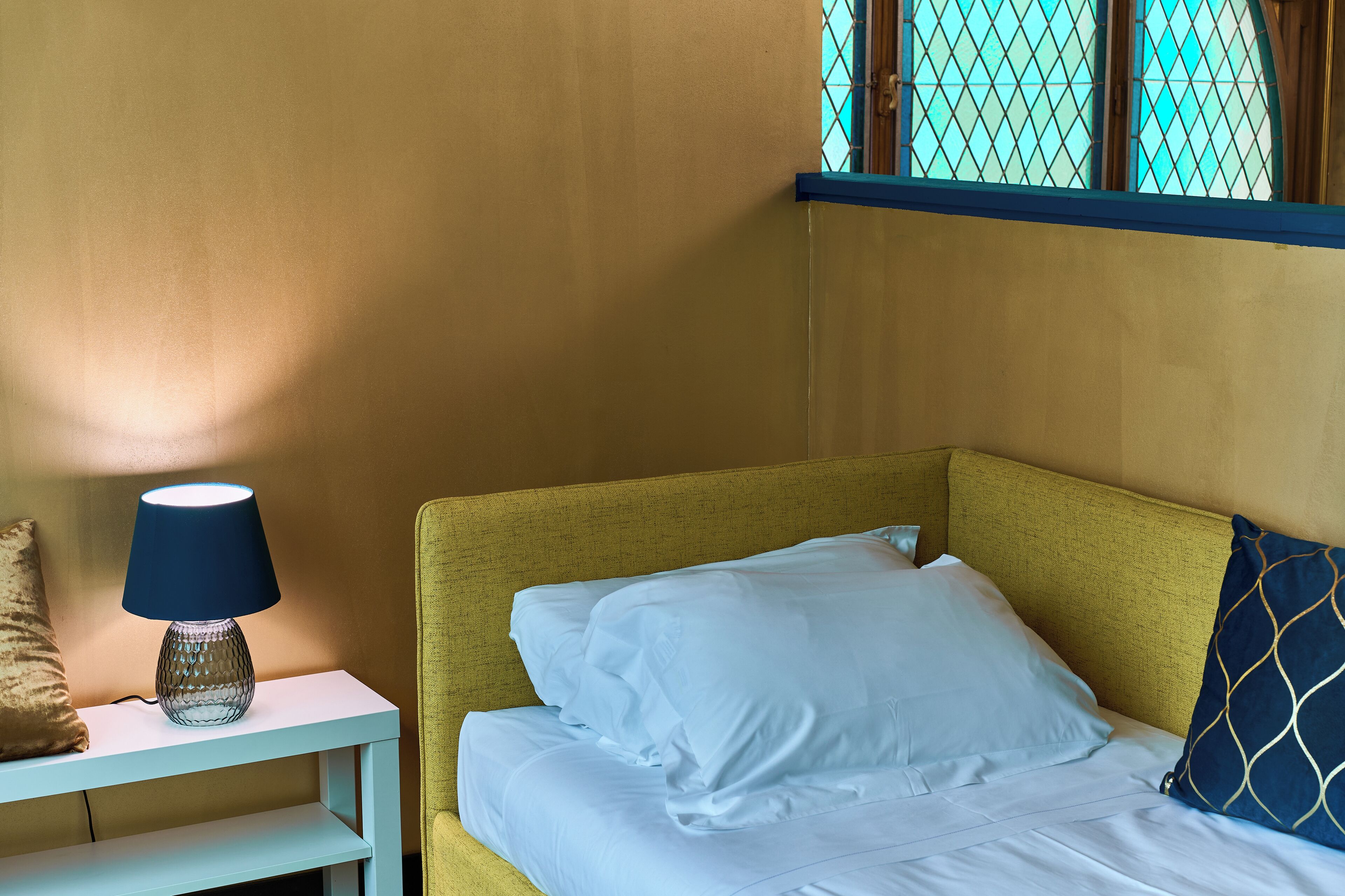 A bed with white bedding and a blue pillow, a bedside table with a lamp, and a window with a blue frame.