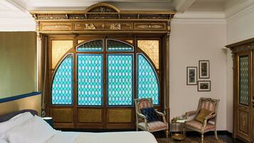 A room with a bed, two chairs, a small table, and a decorative arched window with stained glass.