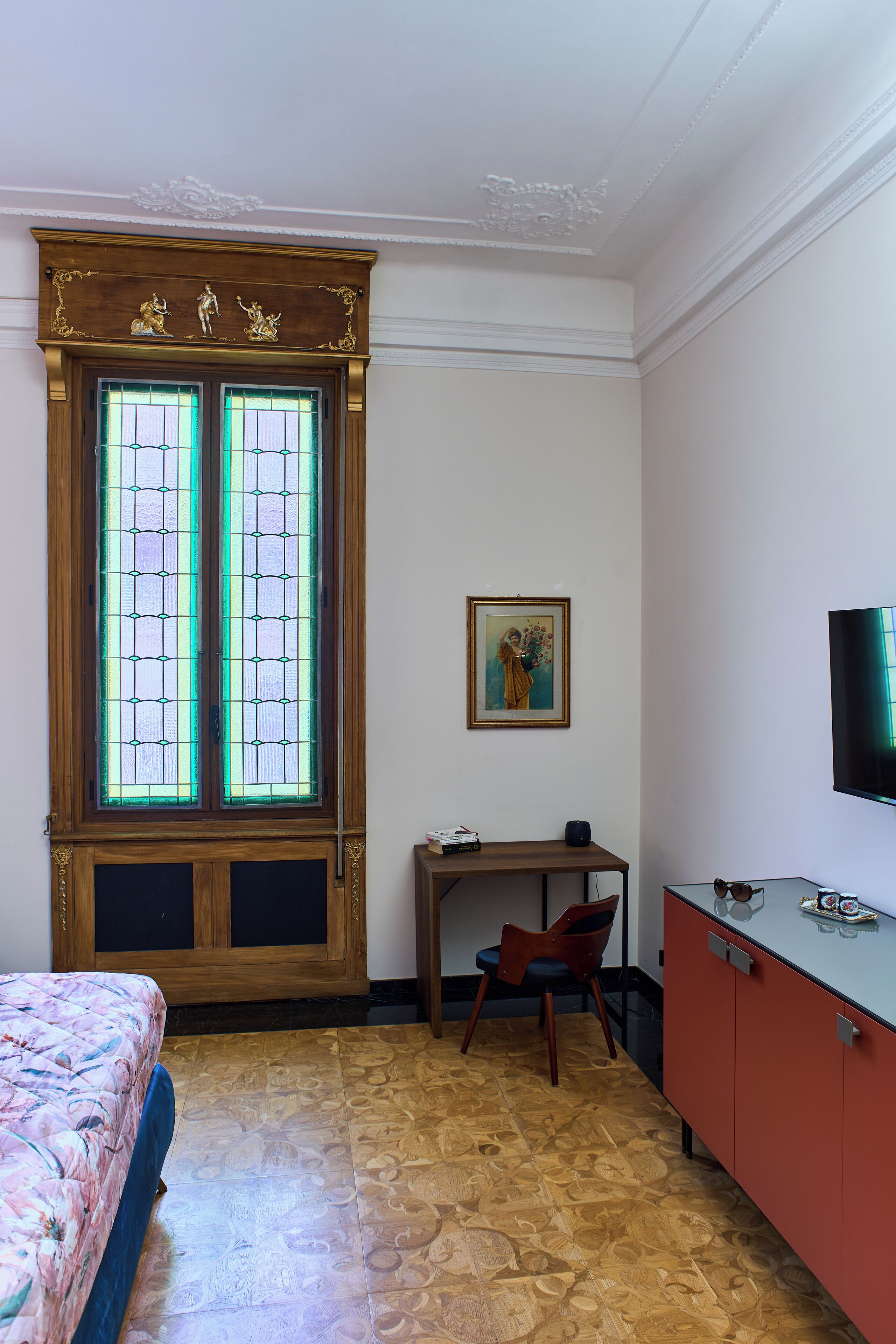 A room with a large wooden door with glass panels, a bed with a floral bedspread, a desk with a chair, a television, and a framed picture on the wall.