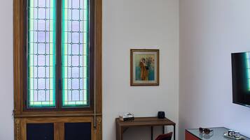 A room with a large wooden door with glass panels, a bed with a floral bedspread, a desk with a chair, a television, and a framed picture on the wall.