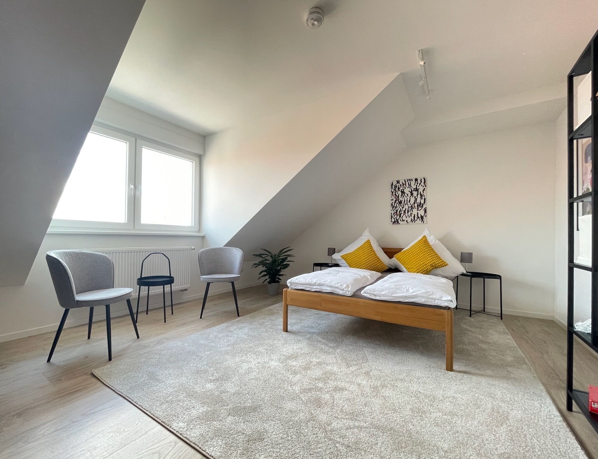 Quarto moderno com teto inclinado, uma cama com almofadas brancas e amarelas, uma poltrona cinza, uma mesa pequena e uma estante.