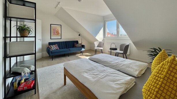 Une chambre de style loft moderne, avec un plafond en pente, une grande fenêtre, un canapé bleu, un lit, une bibliothèque et une plante.