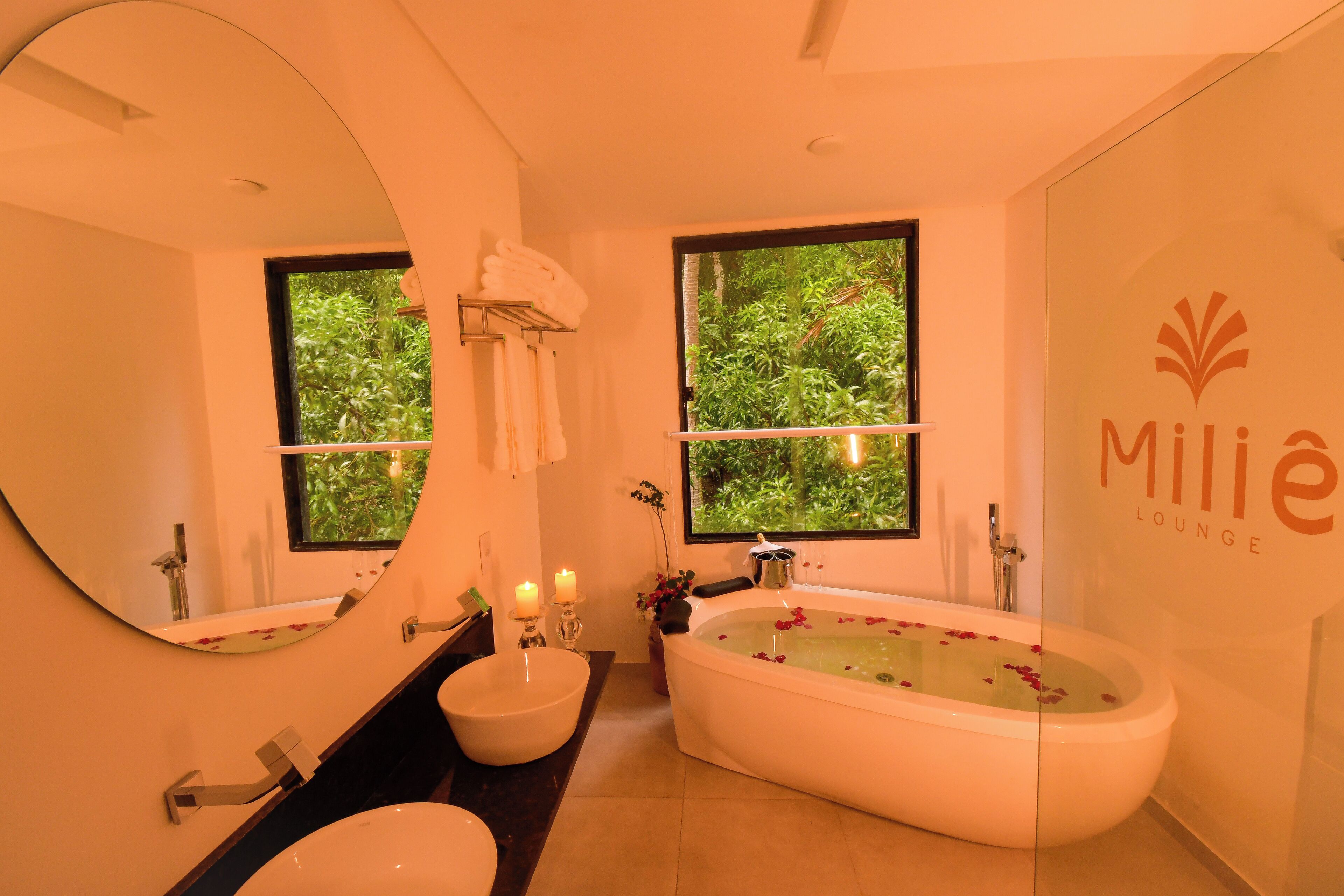 A modern bathroom with a freestanding bathtub, a large mirror, and two windows with greenery visible outside.