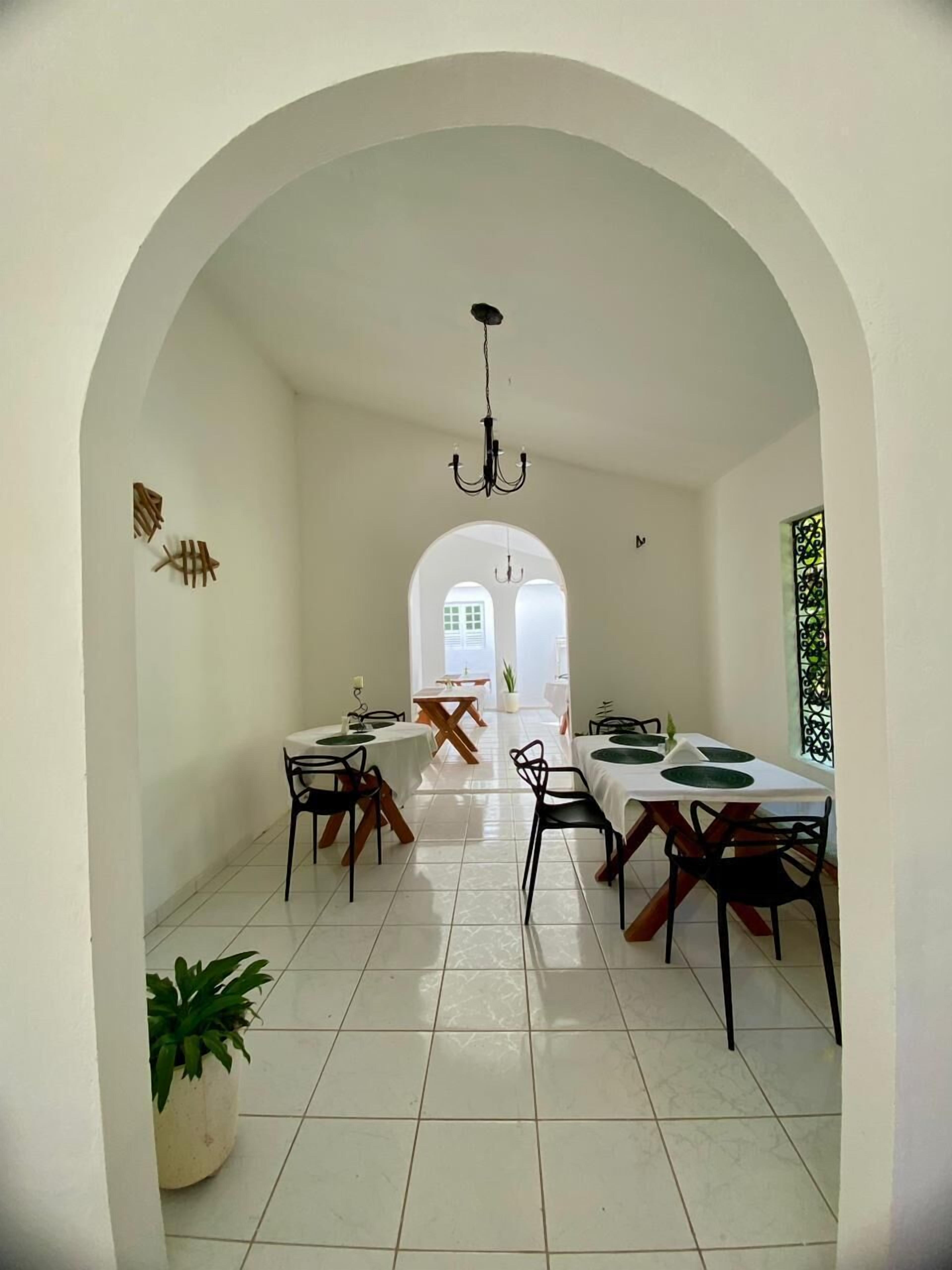 A dining area with a high arched ceiling, white walls, and a tiled floor. There are multiple tables with chairs, a chandelier, and a potted plant.