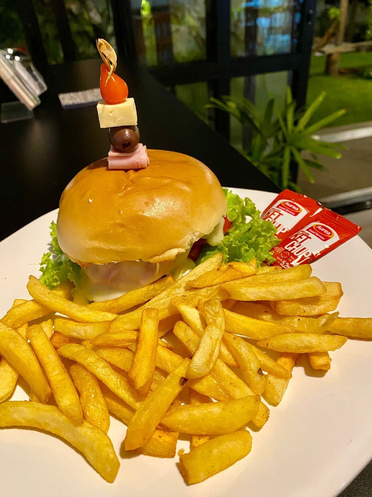 A burger with a toothpick on top, served with French fries and ketchup on a white plate.