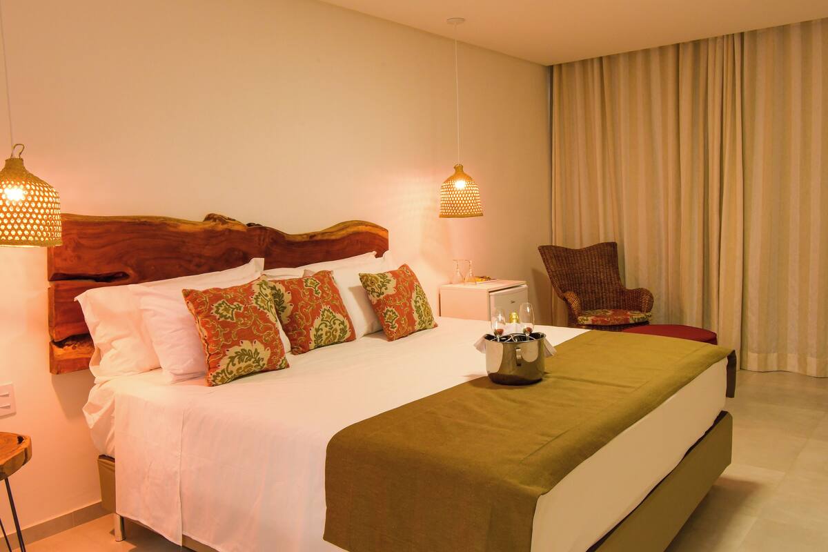 Habitación de hotel con una cama grande, cabecera de madera, dos mesitas de noche, una silla y una ventana con cortinas.