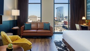 A hotel room with a large bed, a brown leather sofa, a yellow armchair, a small table with a plant, and a view of a cityscape through large windows. - Caption by Hyatt Downtown Nashville (Nashville)