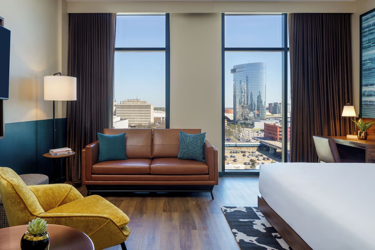 Une chambre d’hôtel avec un grand lit, un canapé en cuir marron, un fauteuil jaune, une petite table avec une plante, et une vue sur le paysage urbain à travers de grandes fenêtres.