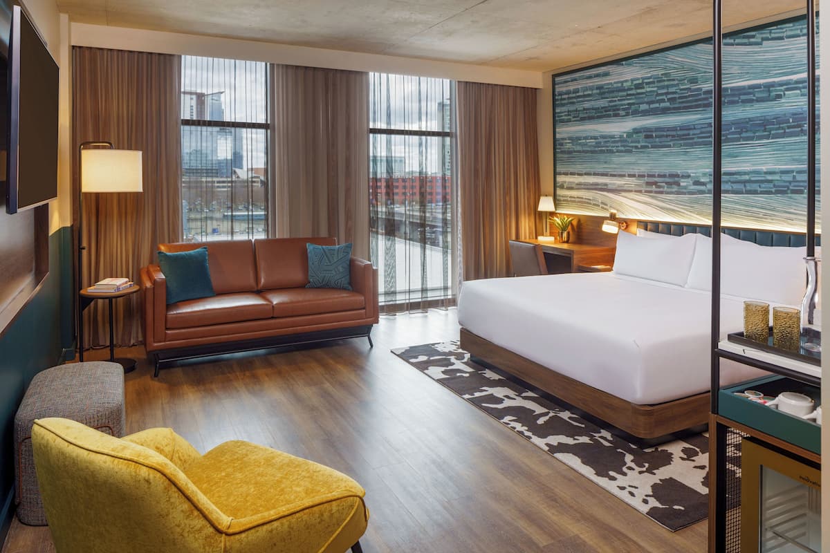 Une chambre d’hôtel avec un grand lit, un canapé, un fauteuil jaune, une petite table et un téléviseur.