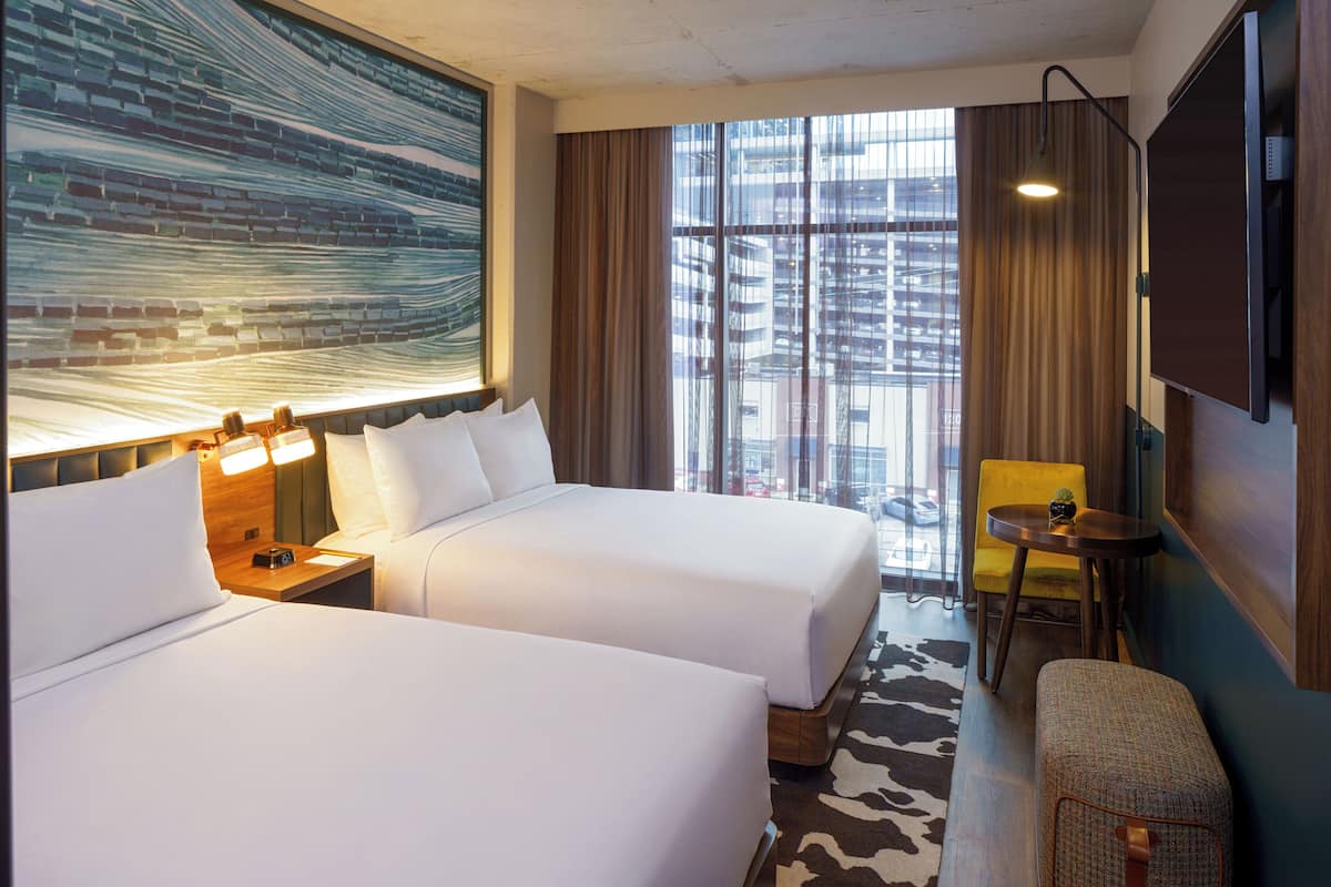 Une chambre d’hôtel avec deux lits, une grande fenêtre avec des rideaux, une petite table, une chaise et un téléviseur fixé au mur.