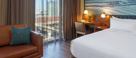 A hotel room with a bed, a desk, a chair, and a sofa. There is a window with curtains, a lamp, and a plant on the desk.