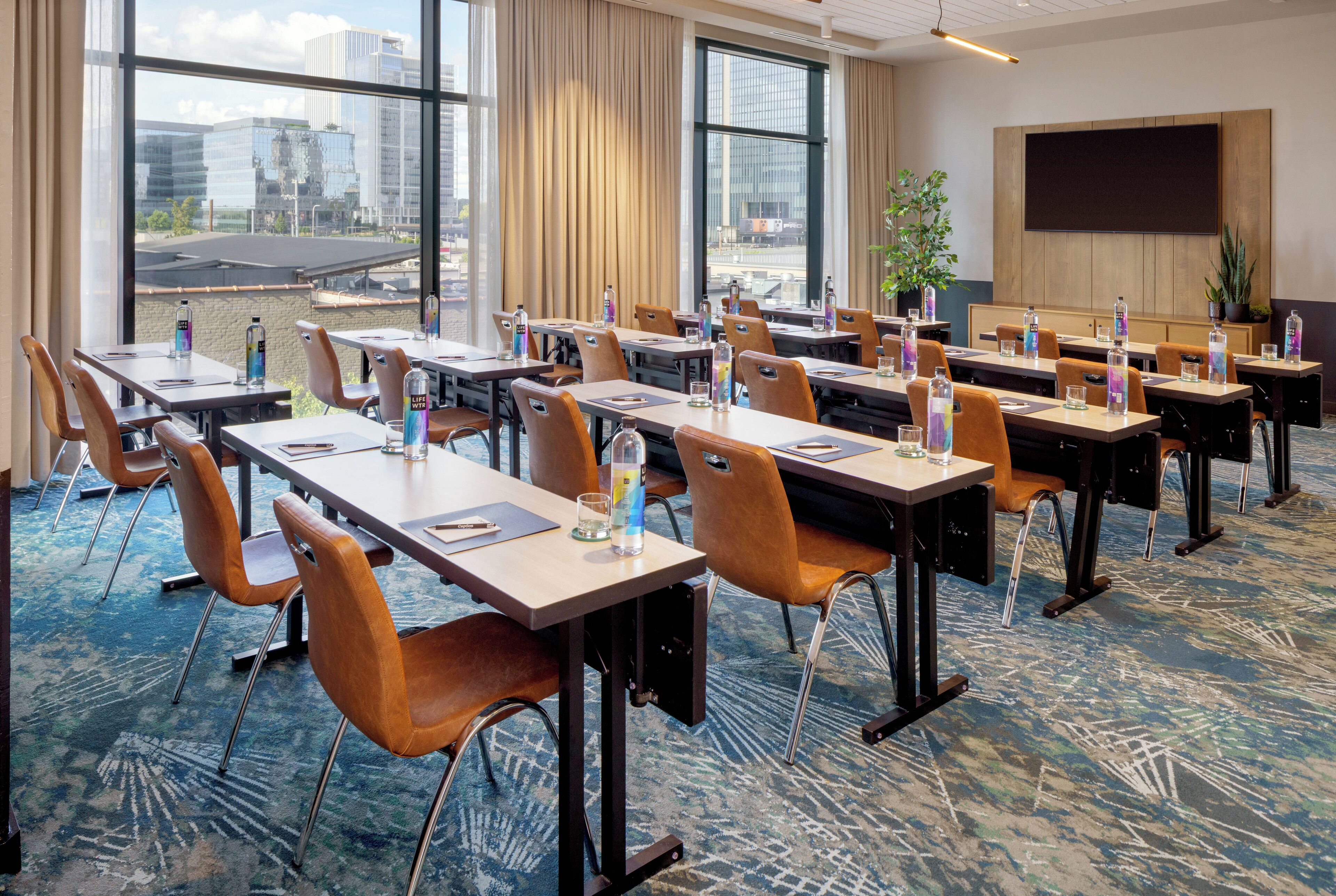 A conference room with tables and chairs, large windows with a city view, a flat-screen TV, and a plant.