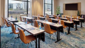 A conference room with tables and chairs, large windows with a city view, a flat-screen TV, and a plant.