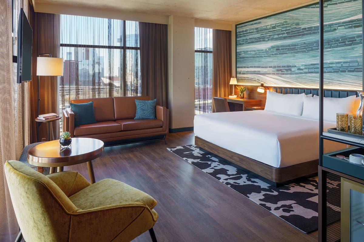 Une chambre d’hôtel avec un lit, un canapé, une chaise, un bureau et une grande fenêtre avec des rideaux.