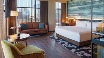 Habitación de hotel con cama, sofá, sillón, escritorio y un amplio ventanal con cortinas.