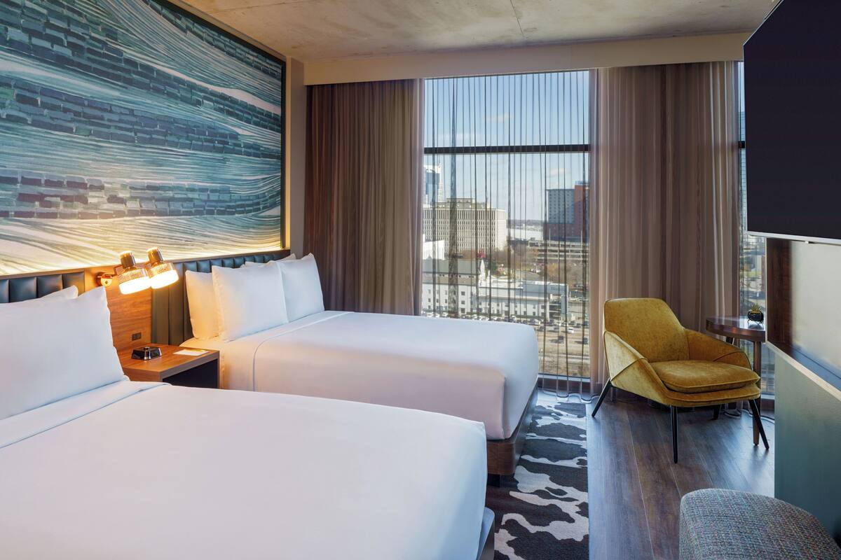 Une chambre d’hôtel avec deux lits, une grande fenêtre avec des rideaux, un fauteuil jaune et un téléviseur fixé au mur.