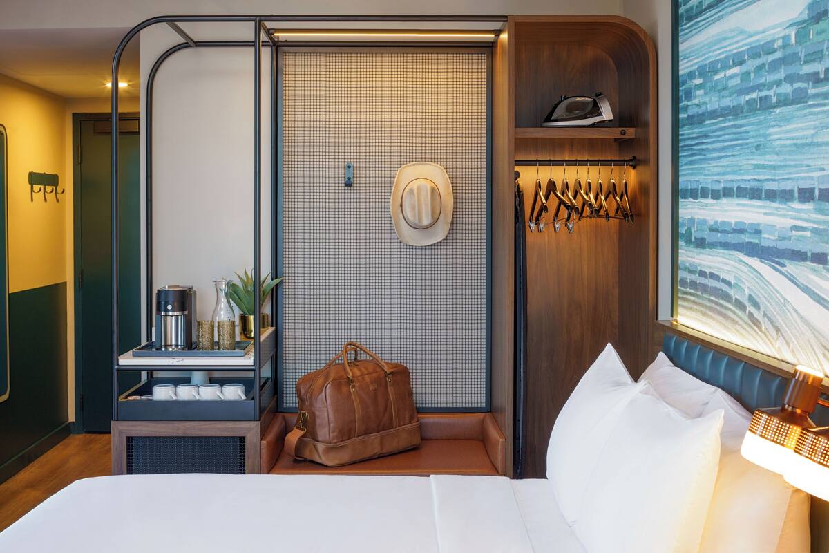 Une chambre d’hôtel avec un lit, une table de chevet avec une lampe, une valise, un chapeau et un porte-manteau.