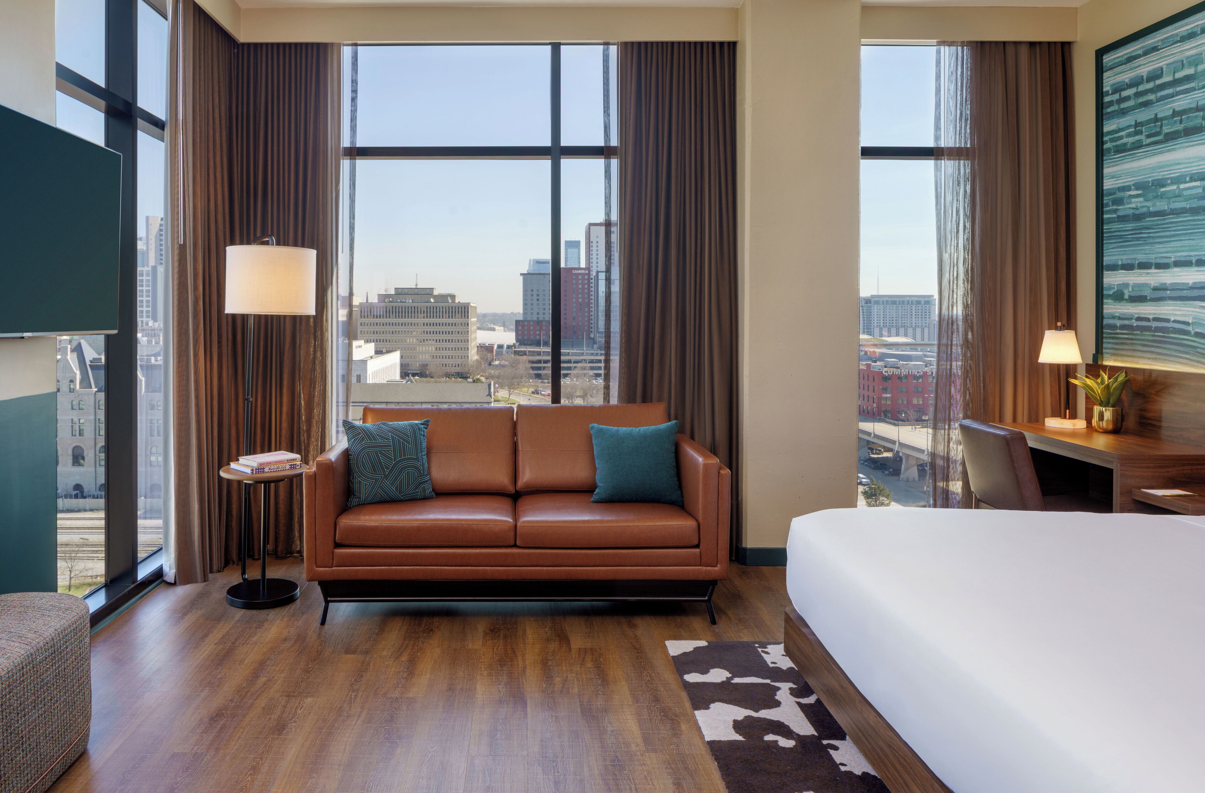 Habitación de hotel con un sofá de cuero marrón, una cama blanca, una mesita redonda pequeña con una lámpara y un amplio ventanal con vistas a la ciudad.