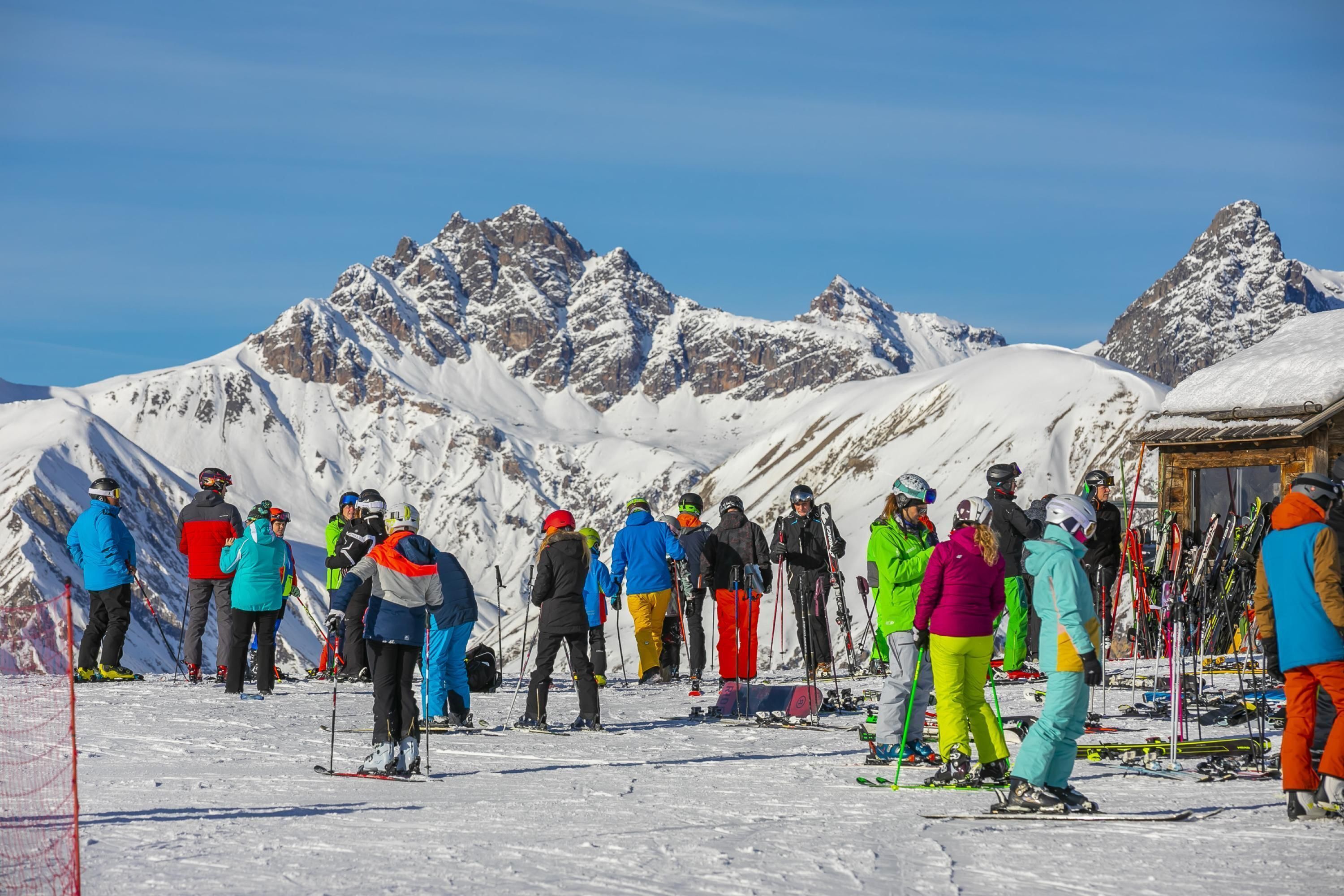 A group of skiers on a snowy mountain slope with a clear sky and snow-covered peaks in the background.