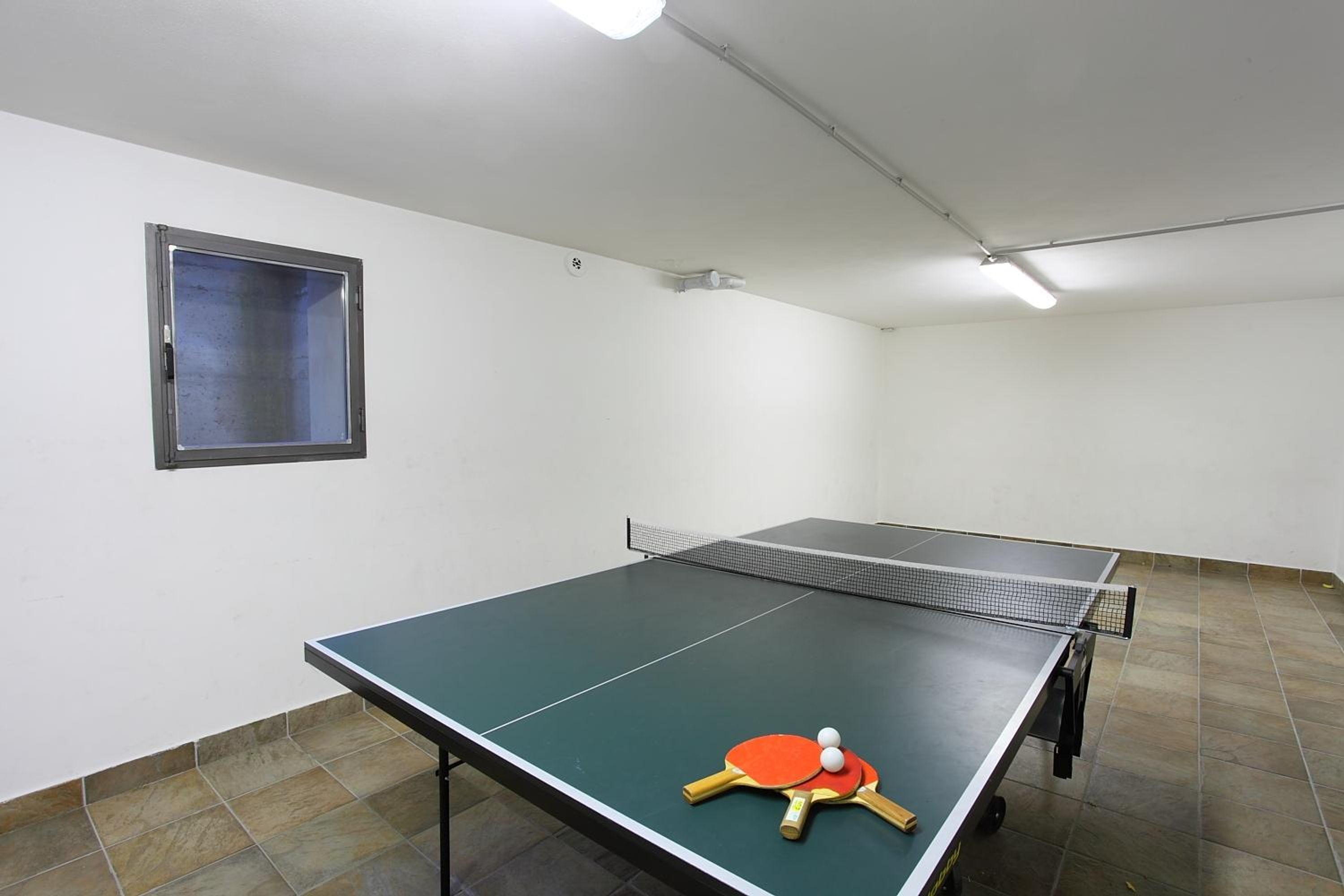 A ping pong table with two paddles and a ball in a room with tiled flooring and a window with a metal frame.