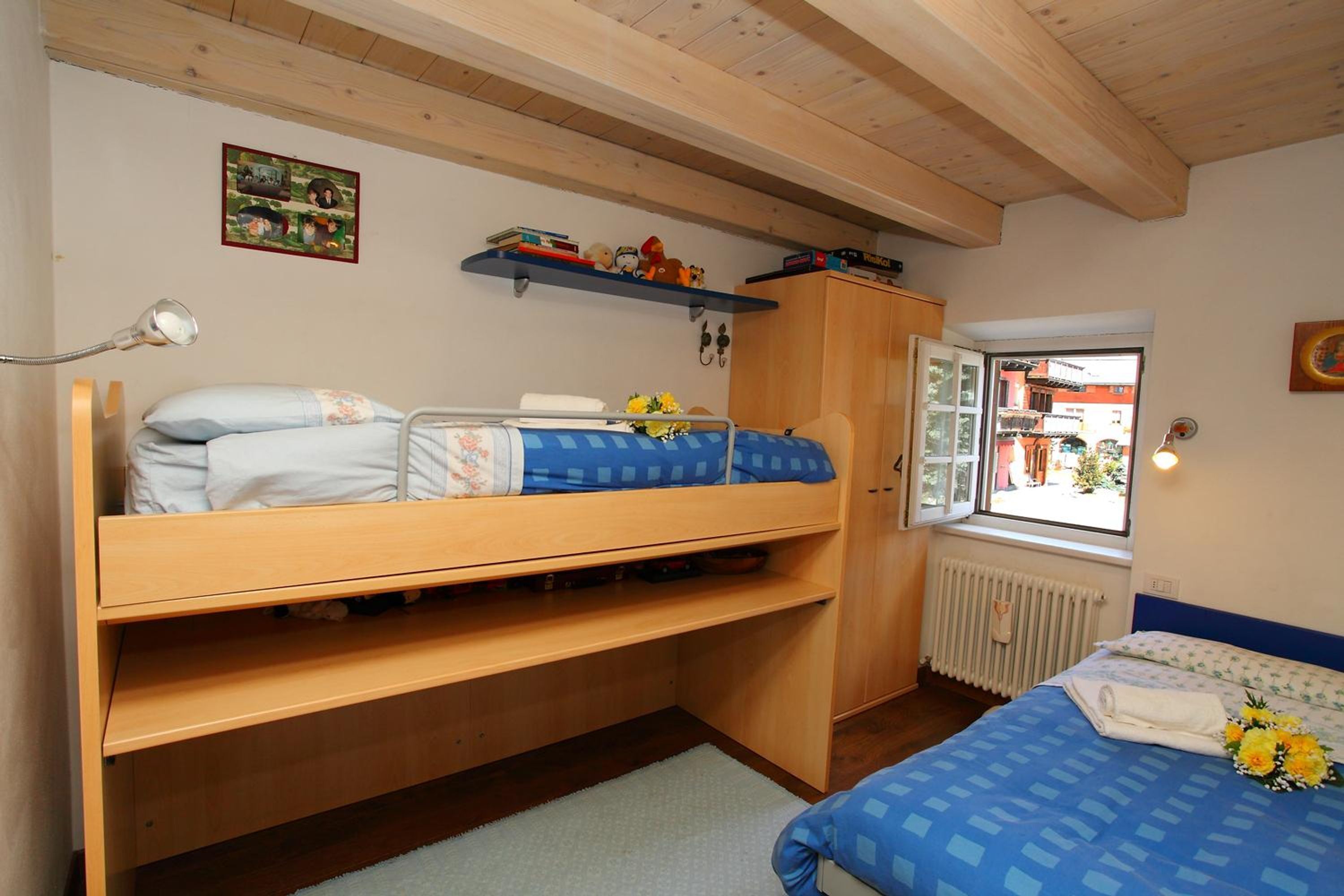 A bedroom with a bunk bed, a desk, a radiator, a window with a view, and a shelf with books and toys.
