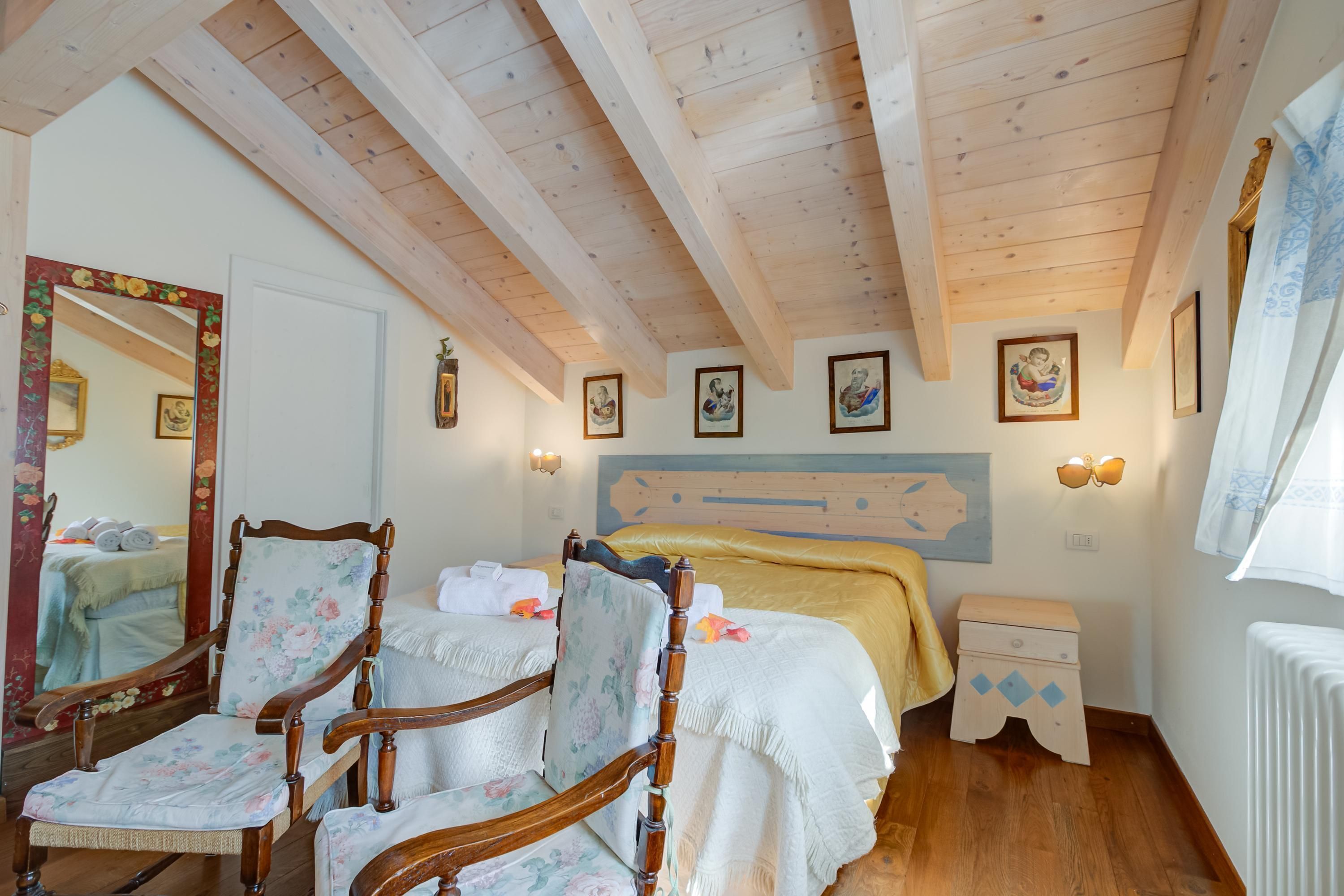 Une chambre avec des poutres apparentes, un lit doté d’une tête de lit jaune, deux chaises en bois recouvertes d’un tissu à motifs floraux, une petite table de chevet en bois et un miroir mural avec un cadre orné de motifs floraux.