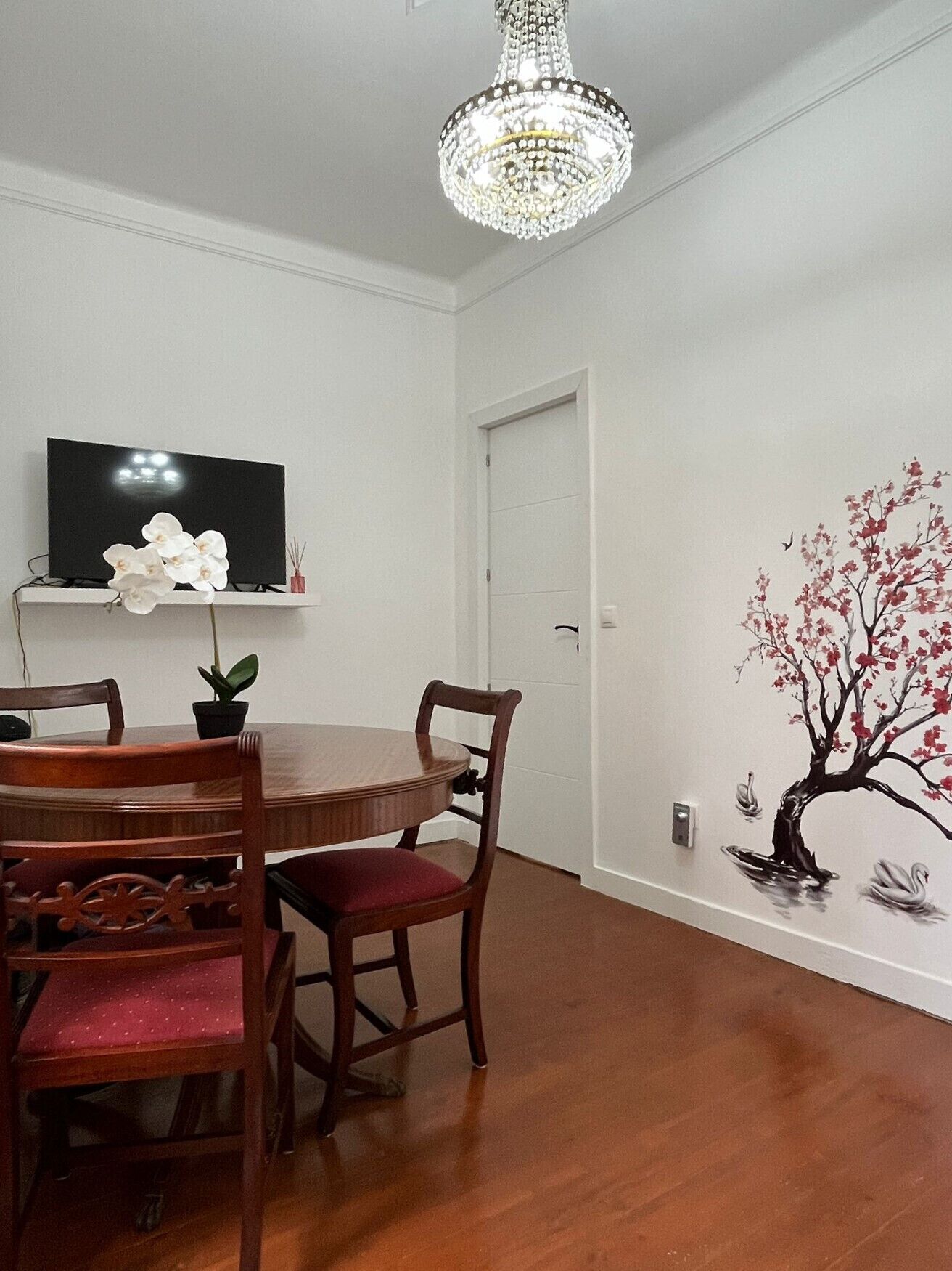 Uma sala de jantar com mesa de madeira, cadeiras, televisão e um candelabro.
