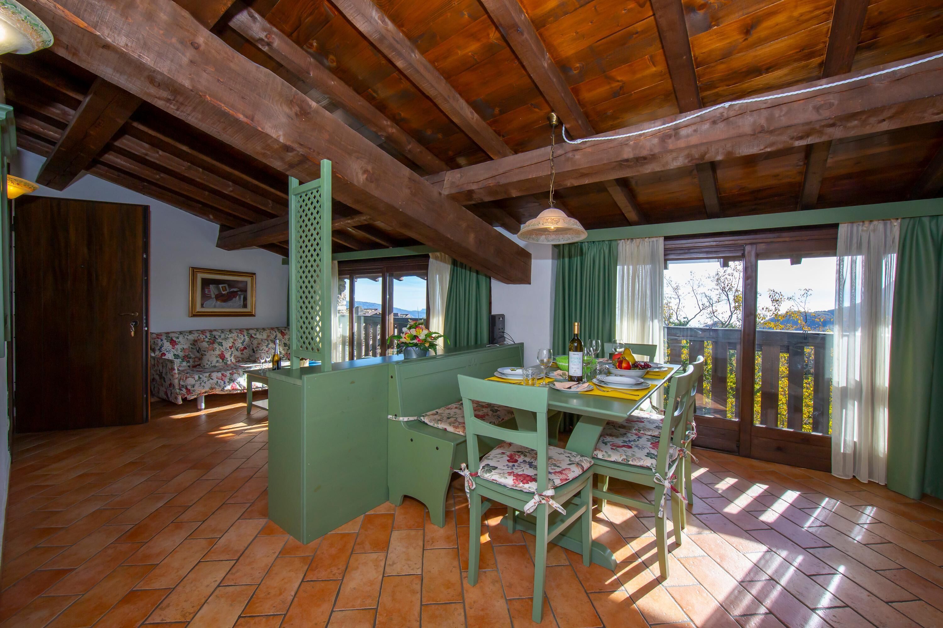 A rustic kitchen with wooden beams, a dining table with chairs, a green island, and a view through the window.