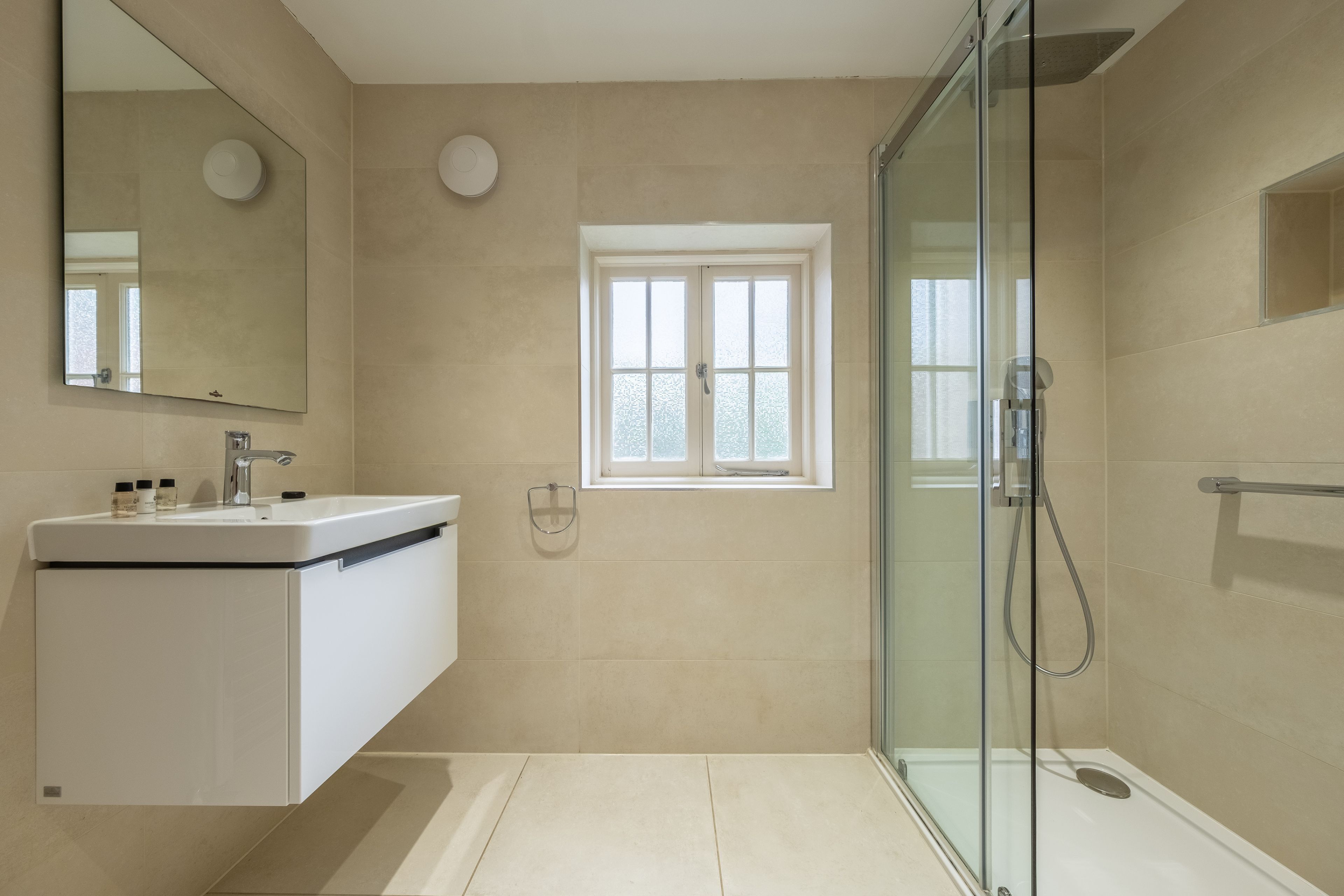 Une salle de bain moderne avec une douche à parois de verre, un lavabo blanc avec un miroir et une fenêtre au-dessus du lavabo.