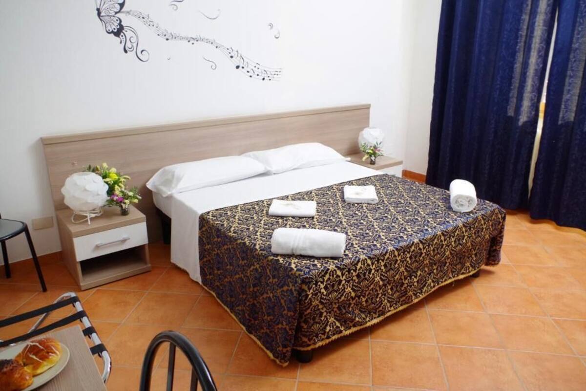 A hotel room with a bed, a nightstand, a chair, a table with a plate of food, and a wall decoration.