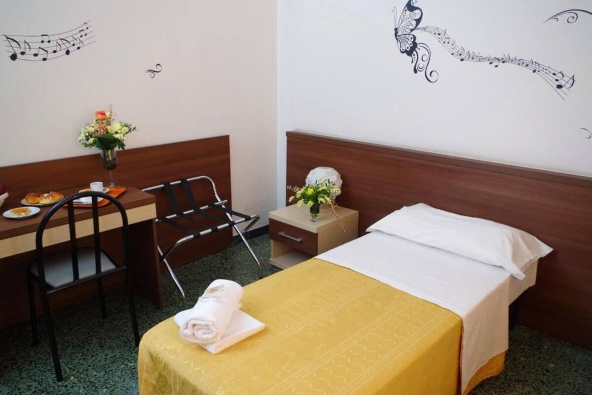 A hotel room with a bed, a desk with a chair, a small table with a vase of flowers, and a wall decoration of a musical note and butterfly.