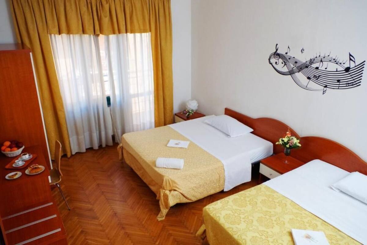A hotel room with two beds, a wooden wardrobe, a dining table with breakfast items, and a wall decoration of musical notes.