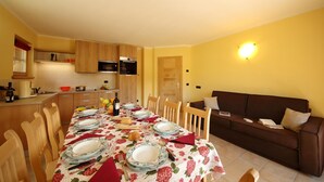 Un comedor con una mesa preparada para cuatro, sillas de madera, un sofá marrón y una cocina al fondo.
