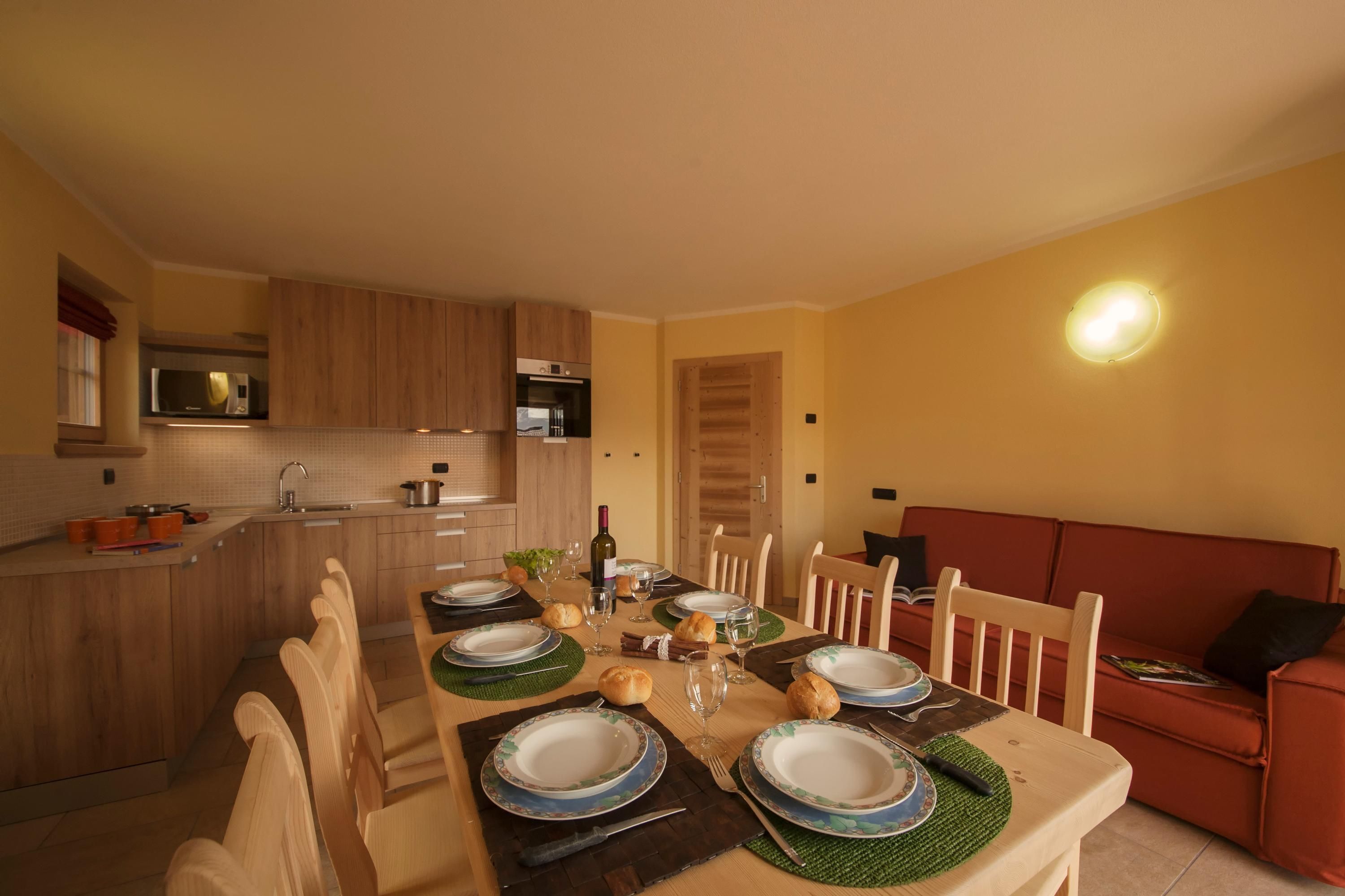 Zona pranzo con tavolo in legno apparecchiata per quattro persone, circondata da sedie, con una cucina sullo sfondo.