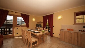 A dining area with a wooden table set for six, a kitchen with wooden cabinets, and a view of mountains through the windows. - Sponda Ski in- Ski out Mt 50 (Livigno)