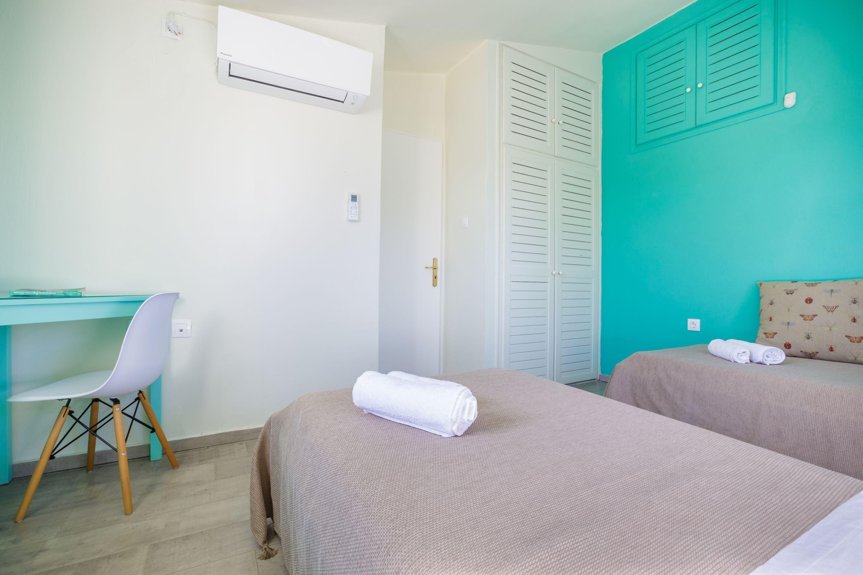 A hotel room with two beds, a desk with a chair, an air conditioner, and a teal-colored wall.