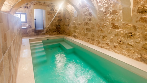 Una piscina interior con paredes de piedra, una fuente de agua azul cristalina, techos abovedados y un pequeño ventanal.