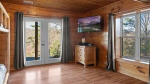 A wooden cabin interior with a wooden floor, a television on the wall, and a window with a view of trees. - Spectacular Secluded Fire Pit Arcade Jacuzzi (Sevierville)
